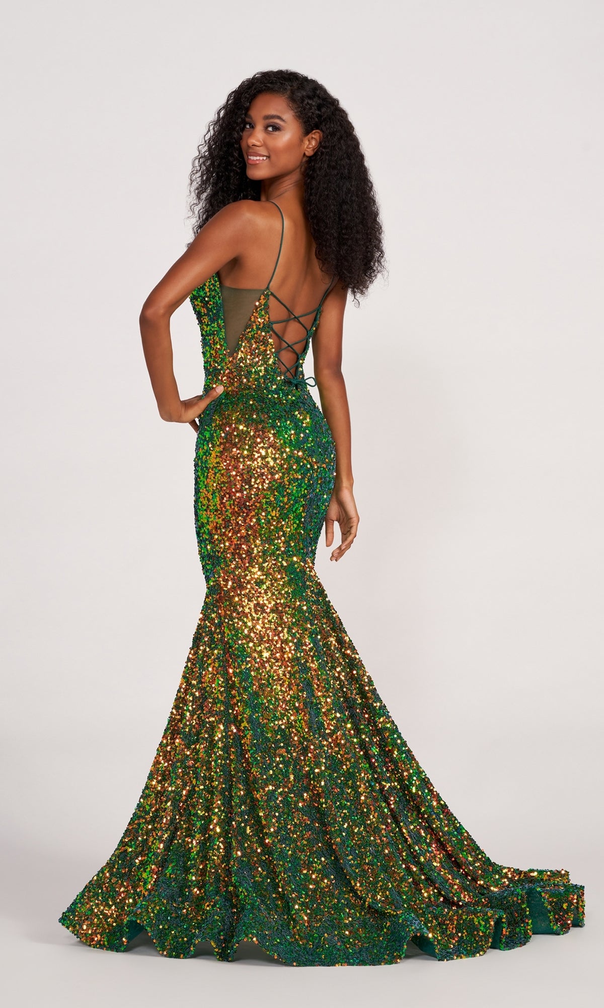 A woman in an Ellie Wilde Long Stretch-Sequin Prom Dress with a shimmering green and gold mermaid silhouette and a strappy lace-up back stands elegantly, gazing over her shoulder.